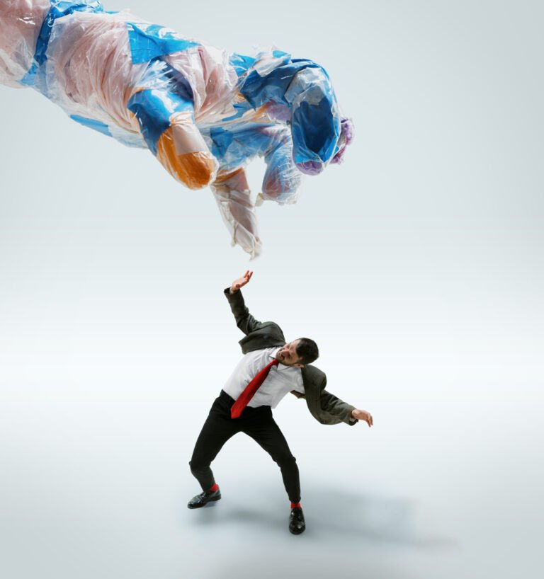 Young caucasian man avoiding big plastic hand on white studio background